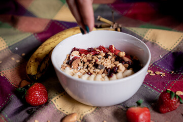 Exquisite oatmeal with banana, strawberries with one hand pouring granola, blueberry, coconut and almonds on top, inside a white plate.
