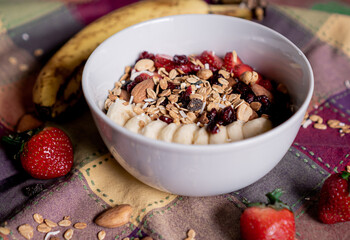 Delicious oatmeal with banana, strawberries, granola, blueberry, coconut and almonds on top, inside a white plate.