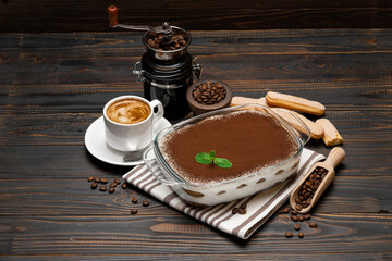 Traditional Italian Tiramisu dessert in glass baking dish, coffee grinder, savoiardi cookies and cup of fresh hot espresso coffee on wooden background