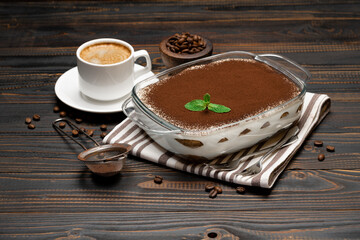 Traditional Italian Tiramisu dessert in glass baking dish and cup of fresh hot espresso coffee on wooden background