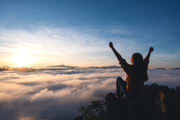 A female traveler raising hands while sitting on the mountain peak, watching sunrise and sea of fog