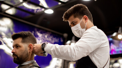 The young and elegant barber with mask cuts the hair of a client with Caucasian features