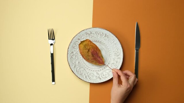 white round plate, knife with fork on brown background, female hand puts dry yellow leaf into plate