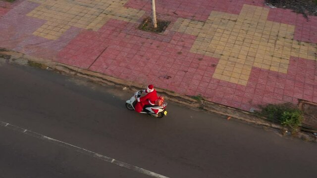 Santa on white motorbike with gift box and gift bag in summer sunset. New year and Christmas content. Boy in Santa costume rides by bike and goes to us. Santa Claus rides a moped, aerial view