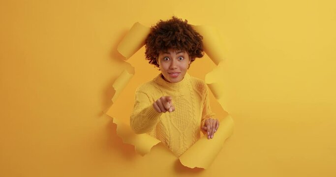 Positive dark skinned young African American woman points directly at camera chooses you picks someone has glad expression dressed in casual yellow jumper stands through torn background. Join me