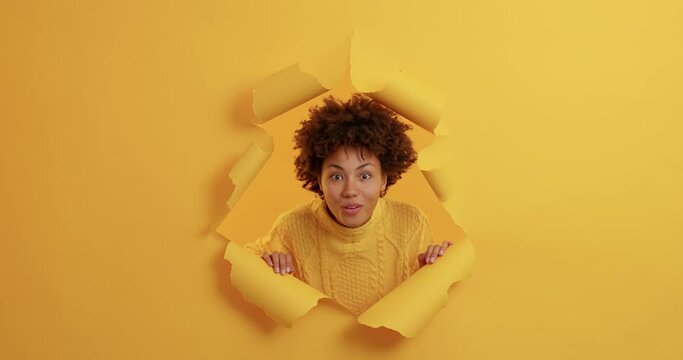 Slow motion of dark skinned woman appearing through paper wall looks surprisingly at camera has unexpected reaction on something breathtaking smiles broadly being in good mood. Yellow background