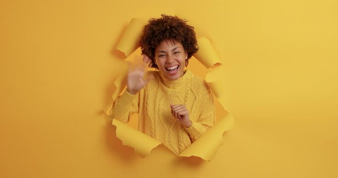 Happy dark skinned young woman waves palm and greets someone has friendly expression stands joyful smiles pleasantly wears casual jumper breaks through yellow paper wall. Hello glad to meet you