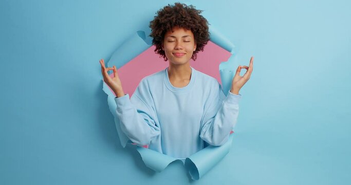 Harmony and peace. Relaxed young African American woman makes mudra gesture practices yoga closes eyes as feels good poses through torn studio wall makes breathing exercise. Blue background.