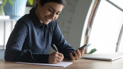 Close up smiling Indian woman holding phone, writing notes, sitting at desk, happy young female...