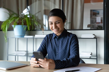 Smiling Indian businesswoman looking at phone screen, sitting in office, happy young female employee executive checking email, workday schedule, browsing mobile device apps, searching information