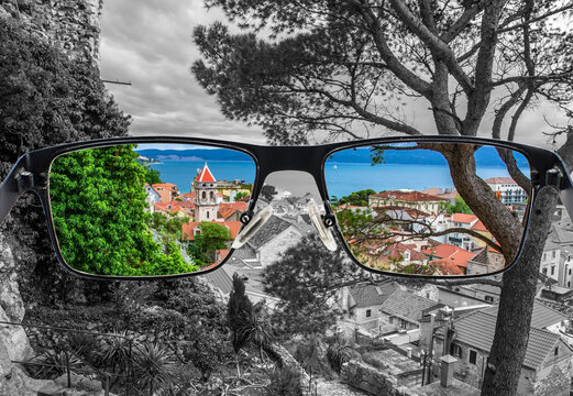 Looking through glasses to colorful view of pine tree,  old city center with red rooftops and blue see surrounded by black and white background. Different world perception. Optimism, hopefulness.