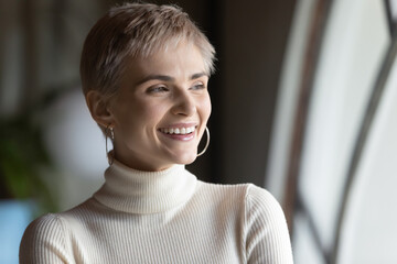 Close up smiling dreamy businesswoman looking to aside through window, visualizing, standing in modern office, happy young female employee intern dreaming about good future, new opportunities