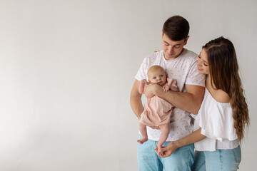 Young caucasian beautiful parents mom and dad hug and kiss their newborn daughter while standing on a white background
