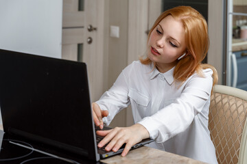 beautiful red-haired girl in a white blouse with a headset uses a laptop at home