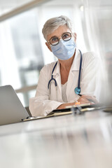 Mature woman doctor with face mask working on laptop