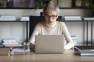 Confident businesswoman wearing glasses working on laptop in office, looking at computer screen,...