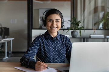 Head shot portrait smiling Indian woman wearing headphones studying online, happy businesswoman intern looking at camera, holding pen, listening to lecture, course, writing notes, watching webinar