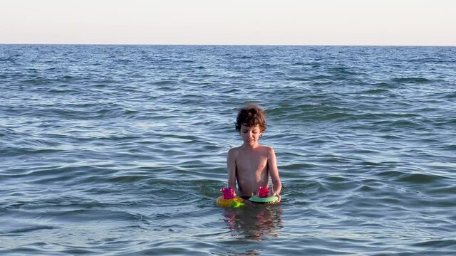 Funny kid playing with inflatable coasters with glasses in sea water. Child swims in the sea at sunset. Little boy on the sea