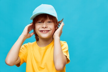Smiling red-haired boy with a cap on his head holds his hands near the face yellow t-shirt