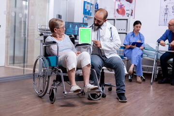 Doctor pointing at green mockup on tablet pc while telling senior woman in wheelchair about her...