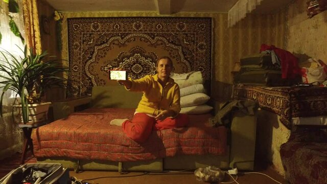 Mature woman is sitting on couch and showing inscriptions on her mobile phone received over the Internet. Atmosphere inside a rural house in village far from civilization