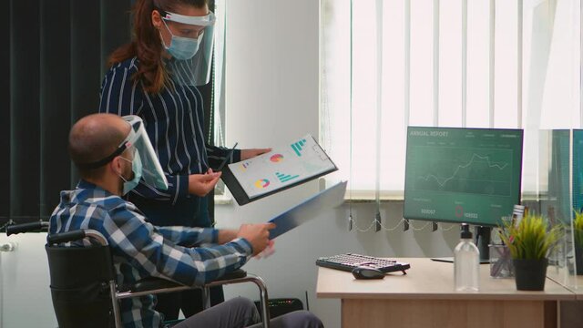Coworkers with masks and visor working in new normal business office, businessman sitting in wheelchair respecting social distance. Team planning project in corporate company during covid-19 pandemic.