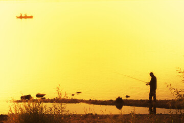 Fisherman with a fishing rod on the river on a beautiful warm yellow-orange sunset background with a boat in the distance