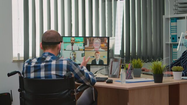 Paralysed manager in wheelchair talking with mask and visor during videocall having online conference in business office. Immobilized freelancer working in financial company respecting social distance