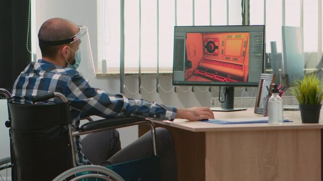 Handicapped game developer sitting in wheelchair with protection mask working at new project from new normal studio office during covid-19 pandemic. Immobilized man respecting social distance.
