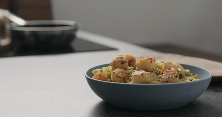 Slow motion add scallops to pesto fusilli in blue bowl on concrete countertop