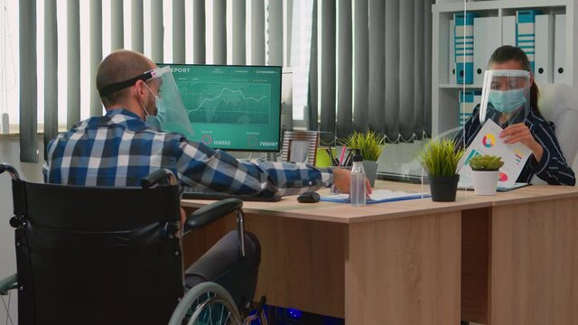 Paralysed financial worker in wheelchair with protection mask working in new normal business office talking with colleague. Immobilized freelancer in corporate company respecting social distance.