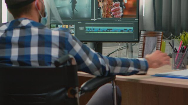 Disabled man videographer with face protection editing video project creating content sitting in wheelchair in new normal office. Blogger working durig global pandemic respecting social distance.