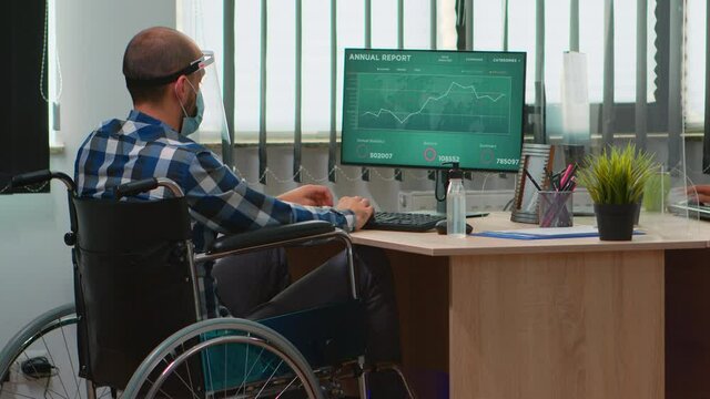 Disabled businessman sitting in wheelchair with protection mask cleaning hands before checking financial data in business modern office. Handicapped freelancer with visor respecting social distance