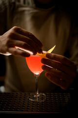 close-up view on man's hands decorates glass with bright red cocktail by fresh lemon peel