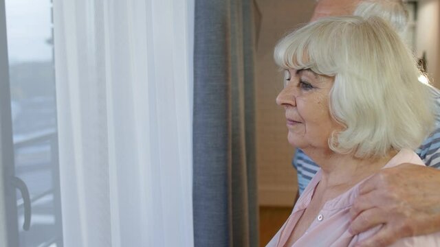 Side view of elegant senior Caucasian grandfather approaches the grandmother from behind. Lovely mature elderly couple is embracing, hugging and looking outside the window of their house. 6k downscale