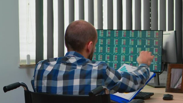 Handicapped businessman working with financial statistics checking graphs sitting immobilized in wheelchair on desk in building office. Paralysed freelacer using modern technology and documents.