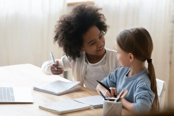 Two diverse little girls doing school tasks, homework together, sitting at table, using laptop, smiling African American and Caucasian sisters studying online, homeschooling concept