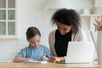 Smiling African American mother helping little adopted Caucasian daughter with school tasks, happy mum and pretty child schoolgirl sitting at table, doing homework, studying together, homeschooling