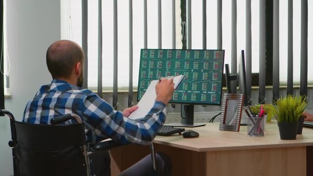 Disabled businessman sitting in wheelchair immobilized checking financial economy data taking notes in business office discussing with colleague. Handicapped freelancer using modern technology.