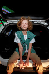 Fototapeta na wymiar An artistic redhead young woman with freckles looking into a camera twisting her face and lips while sitting inside of an opened car trunk with a led screen behind