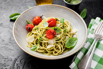 Pasta spaghetti with pesto sauce, grilled tomato and fresh basil leaves in gray bowl on black background.	