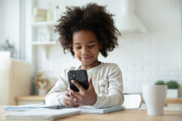 Pretty little African American girl using phone, looking at screen, distracted from studying,...