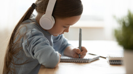 Close up side view little girl wearing headphones writing in notebook, cute child schoolgirl doing...