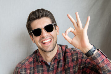 Portrait of a cheerful young dude in black sunglasses with a big smile showing an OK gesture within fingers