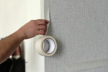 Hand of Man sticks masking adhesive tape on the gray wallpapers on the wall. Roll of adhesive tape. used for painting and preparatory work. Concept of apartment renovations.