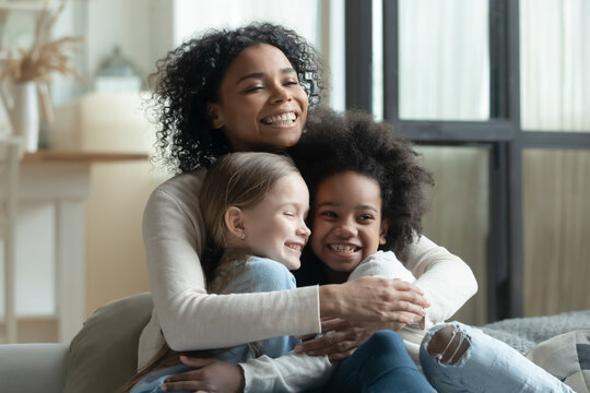 Overjoyed African American woman cuddling with two little daughters, multiracial family enjoying tender moment, sitting on cozy couch at home, loving mum hugging kids, adopted child