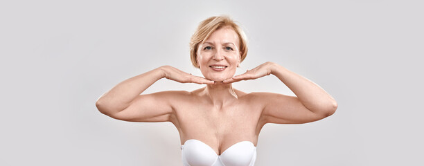 Portrait of beautiful middle aged woman satisfied with skincare treatment. She is holding hands near her face, smiling at camera isolated against grey background. Beauty concept