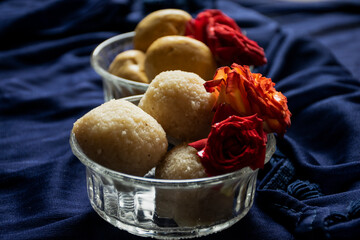 Picture of delicious Rawa laddu decorated in bowl with fresh red roses in a blue background. Rawa laddus made during diwali festival in India.