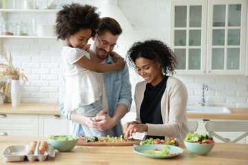 Caring father holding adorable little daughter, standing in kitchen, looking at African American mother cooking salad, shopping vegetables, multiracial family waiting for dinner or breakfast