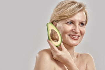 Portait of beautiful middle aged woman smiling aside while holding half of a ripe delicious avocado near her face, posing isolated over grey background